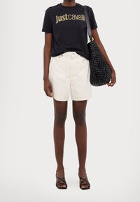 Czarna koszulka z złotym logo "Just Cavalli", zestawiona z jasnymi beżowymi szortami, czarną plecioną torebką i czarnymi sandałami na paski.