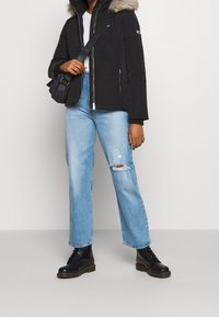 Veste noire avec une capuche en fourrure, fermeture éclair, et poches latérales, associée à un jean bleu clair déchiré et des bottes noires à lacets.