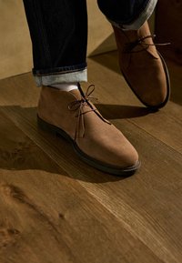 Hnedé semišové chukka čižmy s zaoblenými špičkami, viazaním na šnúrky a členitou podrážkou, kombinované s tmavými džínsami a bielymi ponožkami.