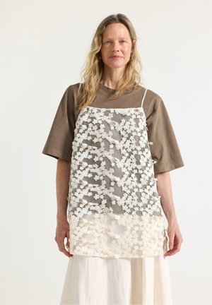 Donna con capelli biondi e lunghi indossa una maglietta marrone oversize sotto un vestito senza maniche in pizzo bianco con motivi floreali, in piedi contro uno sfondo semplice.