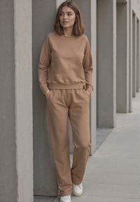 Světle hnědá mikina a odpovídající jogger kalhoty, obojí s hladkým povrchem a uvolněným střihem. Bílé tenisky. Minimalistický design.