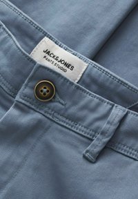 Gros plan sur un pantalon bleu clair avec un bouton et une étiquette blanche visible indiquant « Jack & Jones Pants Studio » à l'intérieur de la ceinture.