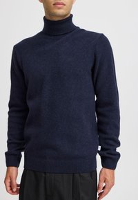Uomo che indossa un dolcevita aderente blu navy scuro con maniche lunghe e polsini a costine, abbinato a pantaloni neri.