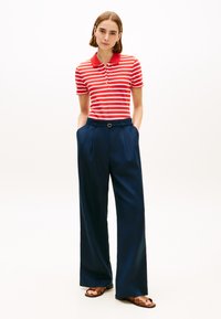 Γυναίκα που στέκεται με τα χέρια στις τσέπες, φορώντας κόκκινη και λευκή ριγέ πόλο μπλούζα, μπλε παντελόνι με φαρδιά πόδια και καφέ σανδάλια σε λευκό φόντο.