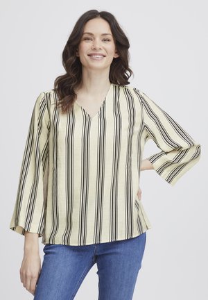 Blouse rayée jaune avec des lignes verticales noires, décolleté en V et manches trois-quarts, fabriquée dans un matériau léger. Associée à un jean bleu.