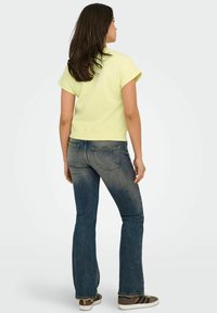 Femme aux longs cheveux noirs portant un haut jaune à manches courtes, un jean bleu délavé et des baskets marron, debout de dos.