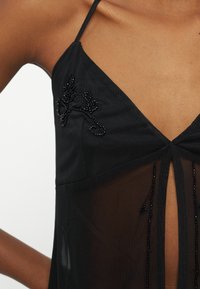 Κοντινό πλάνο ενός ατόμου που φοράει μαύρο διάφανο φόρεμα με λεπτές τιράντες και κεντήματα με χάντρες σε σχήμα λουλουδιών κοντά στο λαιμό.