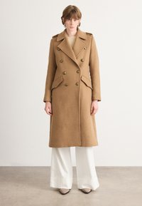 Palton dublu în camel din lână cu țesătură texturată, nasturi mari și buzunare laterale, purtat cu pantaloni albi cu picioare ample.