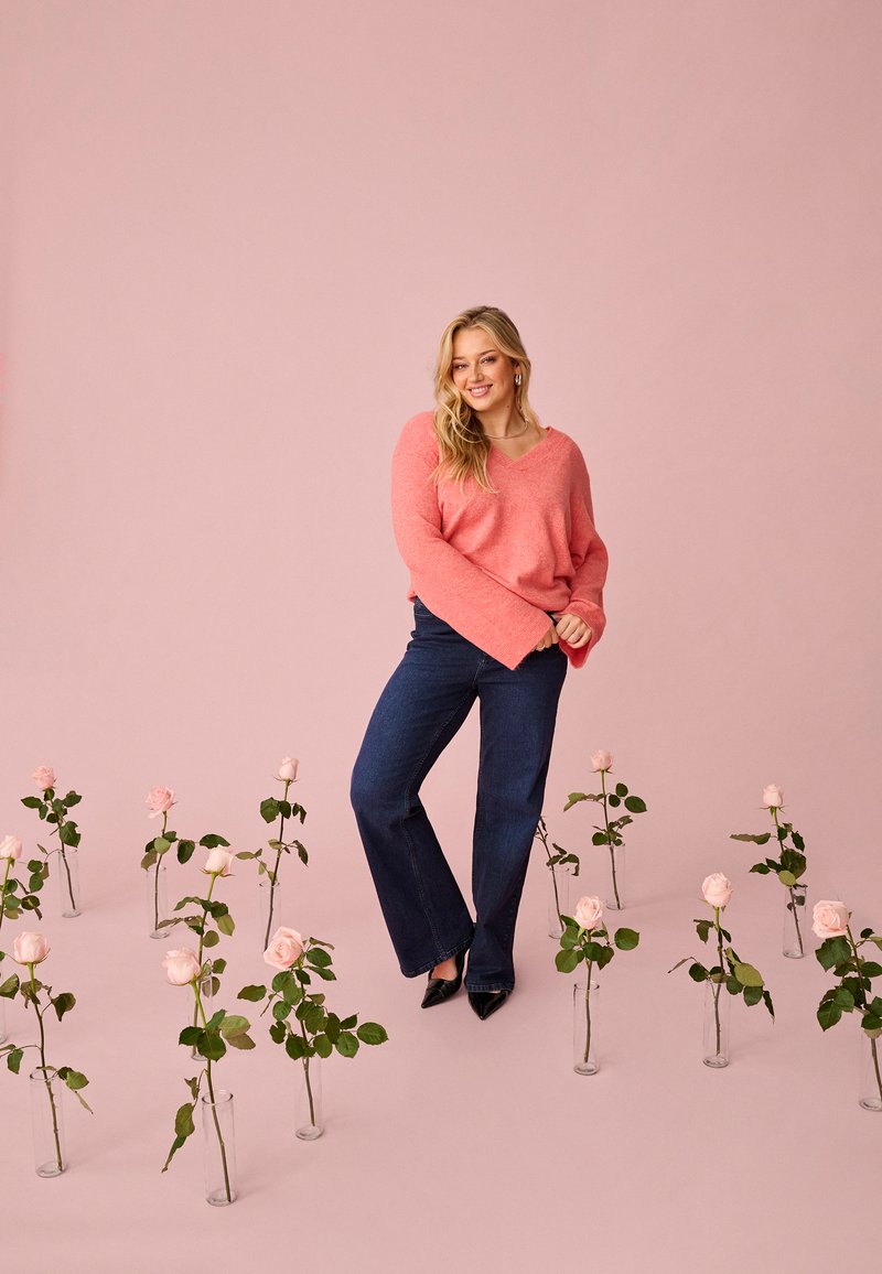 Mladá žena v korálovom svetri a tmavých džínsoch stojí medzi sklenenými vázami s ružovými ružami na jemnom ružovom pozadí.