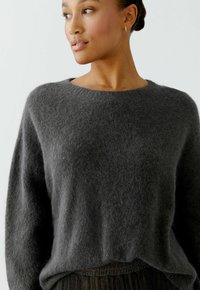 Dark grey, fuzzy sweater with a round neckline and loose fit, featuring subtle texture. Worn over a pleated, darker garment.