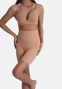 Donna che indossa un reggiseno modellante senza cuciture color carne e pantaloni corti modellanti a vita alta, in piedi con una gamba incrociata sull'altra.