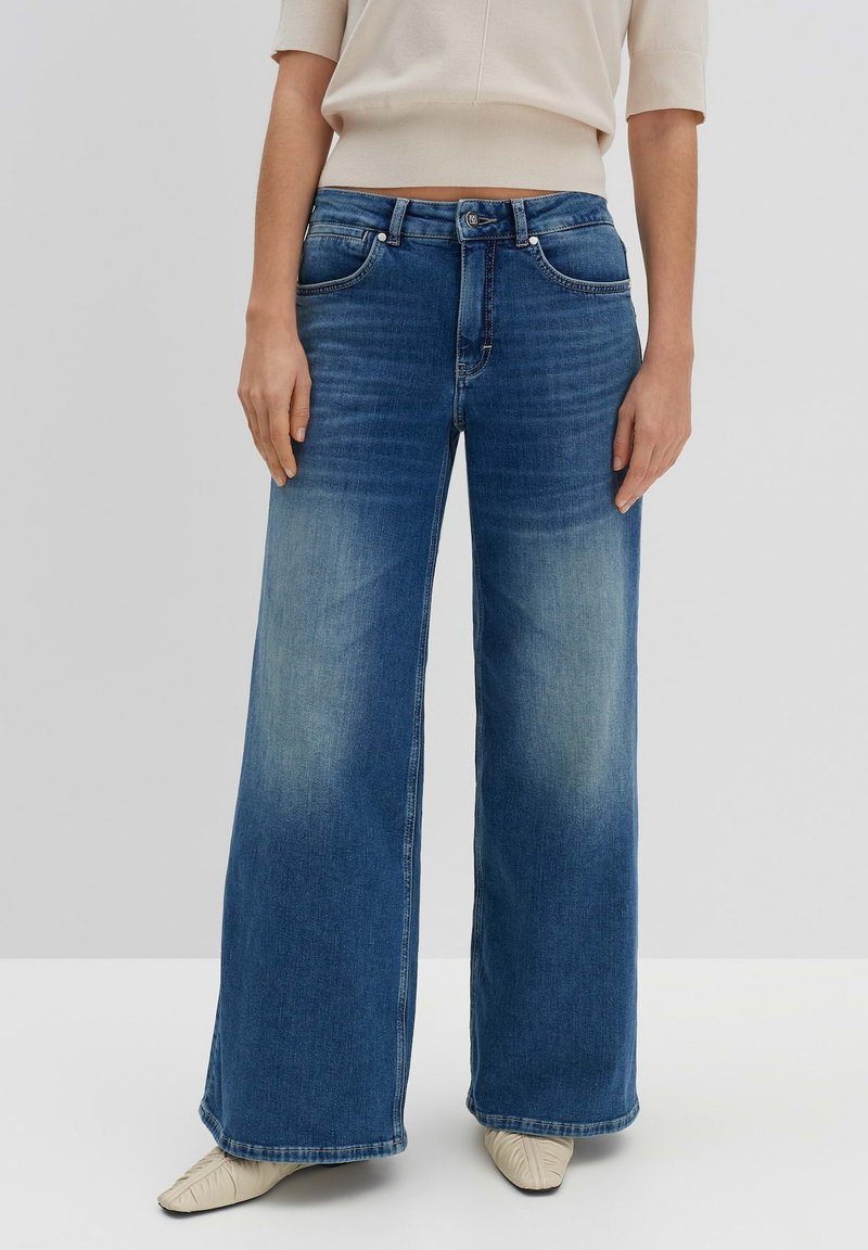 Jean en denim bleu à jambe large avec une finition délavée, design à cinq poches et fermeture à boutons, associé à un haut de couleur claire.