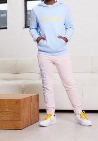 Ljusblå hoodie med gul "LACOSTE"-logotyp, kombinerad med rosa sweatpants med "LACOSTE" tryckt i blått. Vita sneakers med gula skosnören.