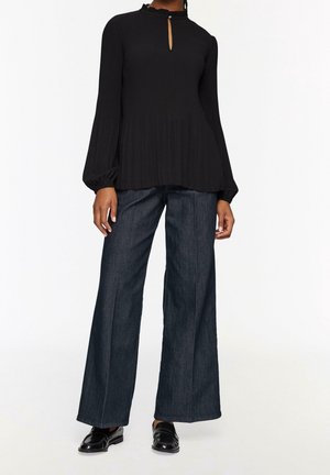 Femme portant un chemisier noir à manches longues avec une encolure en forme de trou de serrure, un jean large foncé et des mocassins noirs vernis sur un fond uni.