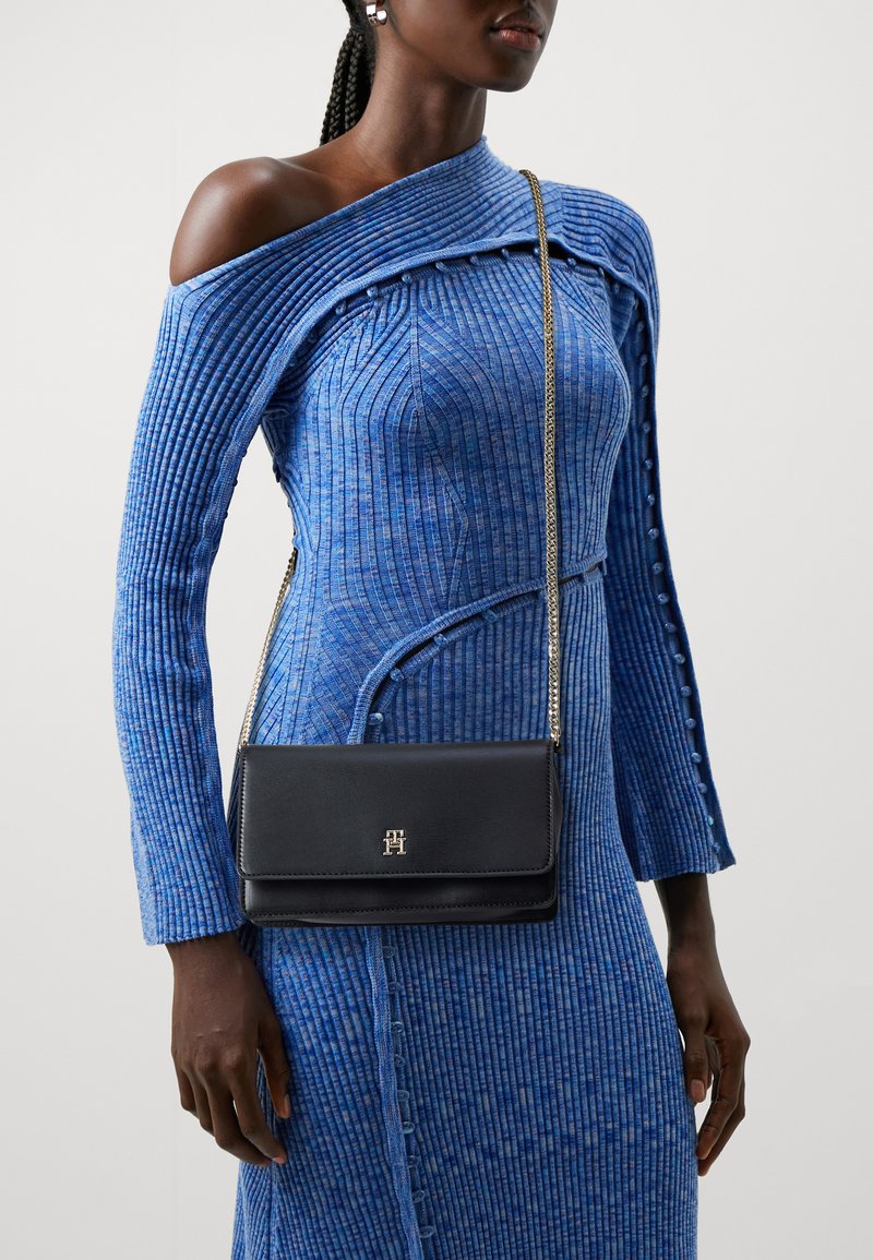 Abito blu a coste con design off-shoulder e dettagli con bottoni, abbinato a una borsa a tracolla in pelle nera con una tracolla in oro e logo.