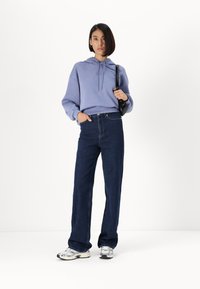 Lilla hettegenser med snøring, kombinert med høyt liv blå jeans og sølvsko. Kvinne står med den ene hånden i lommen.