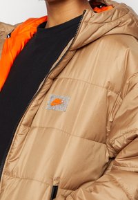 Brun puffjacka med en blank yta, orangerand och en tydlig etikett med en liten orange logotyp på vänster bröst.