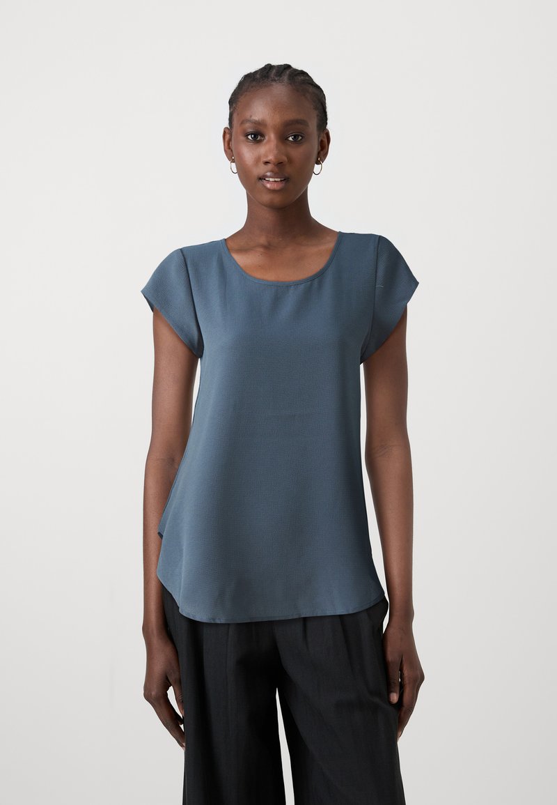 Short-sleeve, blue-gray top made of smooth fabric. Features a round neckline and a curved hem. Paired with black, wide-legged trousers.