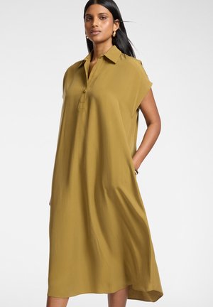 Vrouw met lang zwart haar, gekleed in een mosterdgele, mouwloze midi-jurk met kraag en zijzakken, staand tegen een witte achtergrond.