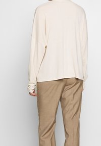 Krämsfärgad långärmad topp med sänkta axlar, rundad halsringning och avslappnad passform. Sammanställd med bruna raka byxor med en subtil textur.