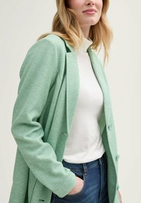 Blazer texturizado en verde menta con solapa muesca, cierre de un botón y dos bolsillos frontales, usado sobre una blusa blanca y jeans azules.