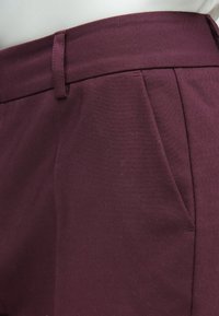 Pantalon sur mesure bordeaux en tissu lisse, doté d'une ceinture plate, de poches latérales et de plis discrets.