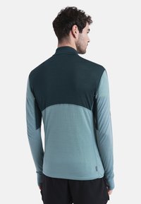 Camiseta deportiva de manga larga en degradado de teal, con una sección superior en verde oscuro, tejido texturizado y costuras laterales abiertas para ventilación.