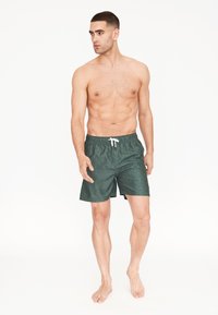 Vihreät kuvioidut uimaportit, joissa on joustava vyötärö, narusäätö ja rento malli, joita käyttää alastomasti rintamuksesta malli, joka seisoo paljain jaloin.