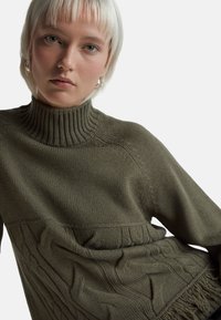 Olivově zelený pletený svetr s vysokým žebrovaným výstřihem, texturovanými panely a třásněmi na dolním lemu. Měkký materiál a volný střih.