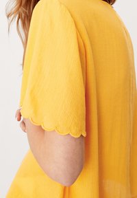 Top giallo con tessuto testurizzato, caratterizzato da bordi delle maniche a scallop e una vestibilità rilassata. Il design include leggere righe verticali.