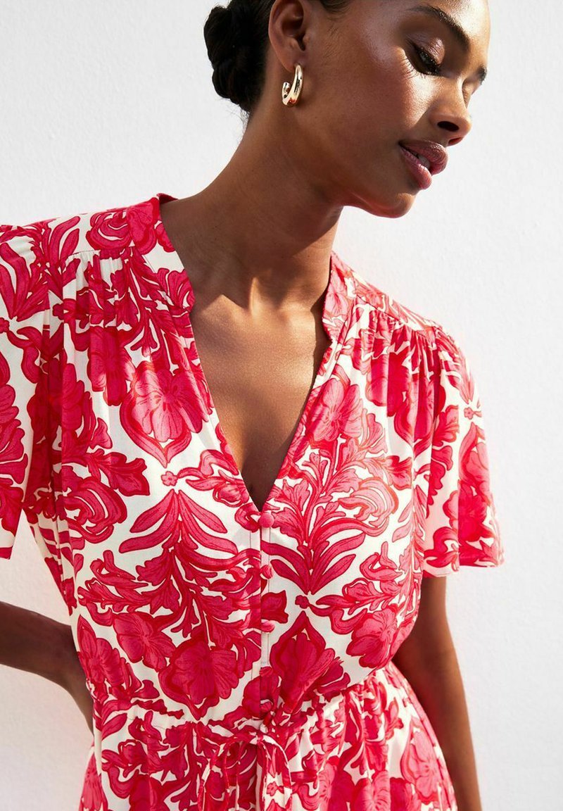 Robe à imprimé floral rouge avec un décolleté en V, des manches courtes et une taille nouée. Le tissu est léger, avec un fond blanc contrastant. Boucles d'oreilles en or.