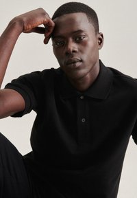 Polo shirt nera con un design classico, maniche corte, patta con tre bottoni e tessuto testurizzato. Il modello ha un'espressione neutra.