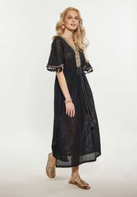 Rochie maxi din dantelă neagră cu guler brodat, mâneci scurte și margini cu franjuri. Asortată cu sandale aurii cu barete.