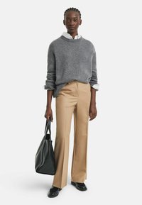 Donna che indossa un maglione grigio sopra una camicia bianca, pantaloni beige, scarpe nere, tiene una grande borsa nera testurizzata, in piedi su uno sfondo bianco.