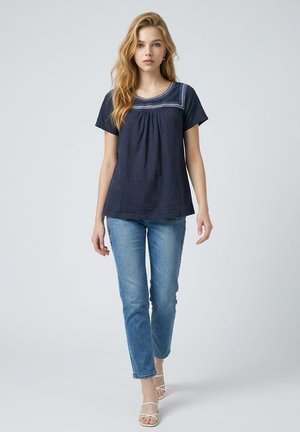 Joven mujer con pelo largo y rubio que lleva una camiseta azul marino de manga corta con cuello bordado, vaqueros azul claro y sandalias blancas con tiras.