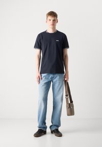 Jūrų aukso medvilnės marškinėliai su baltais akcentais, šviesiai mėlyni džinsai, juodi sportbačiai ir mažas rudas per petį nešiojamas krepšys su peties dirželiu.