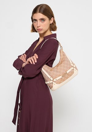 Donna in abito color borgogna che porta una borsa a tracolla beige con motivo e manici in pelle bianca, in piedi davanti a uno sfondo chiaro e uniforme.