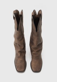 Botas de tornozelo em pele castanha com um design folgado, bicos pontiagudos e uma textura suave. Apresentam duas abas em laço para facilitar o uso.