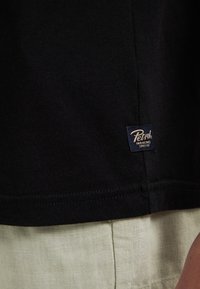 Črna bombažna majica s strukturirano površino in majhnim modrim oznako z logotipom "Petrol" na robu, v kombinaciji s svetlo zelenimi lanenimi hlačami.