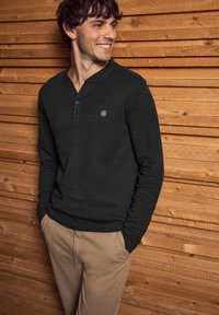 Svart långärmad henley-tröja med tre knappar och en liten logotyp-lapp, tillsammans med beige byxor framför en träbakgrund.