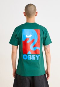 Tricou din bumbac verde, cu un grafic mare multicolor pe spate, ce prezintă forme abstracte și cuvântul "OBEY" în litere albastre în partea de jos.