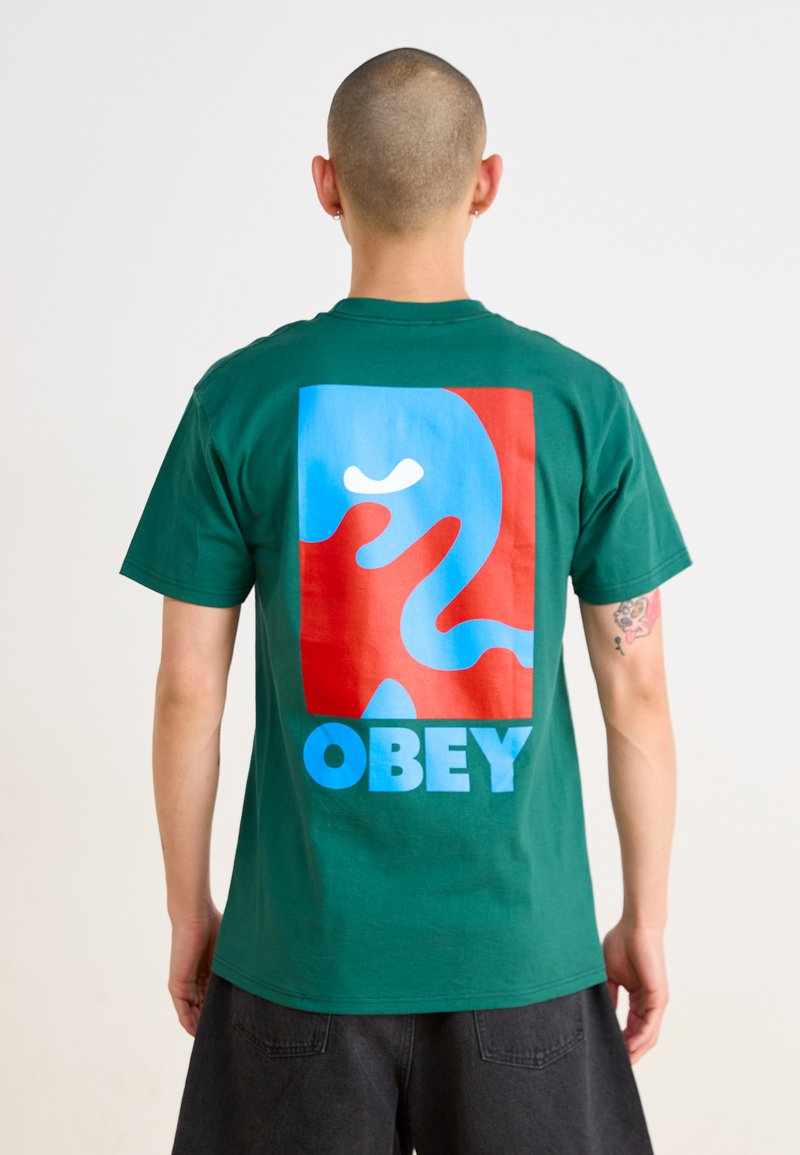 Tricou din bumbac verde, cu un grafic mare multicolor pe spate, ce prezintă forme abstracte și cuvântul "OBEY" în litere albastre în partea de jos.