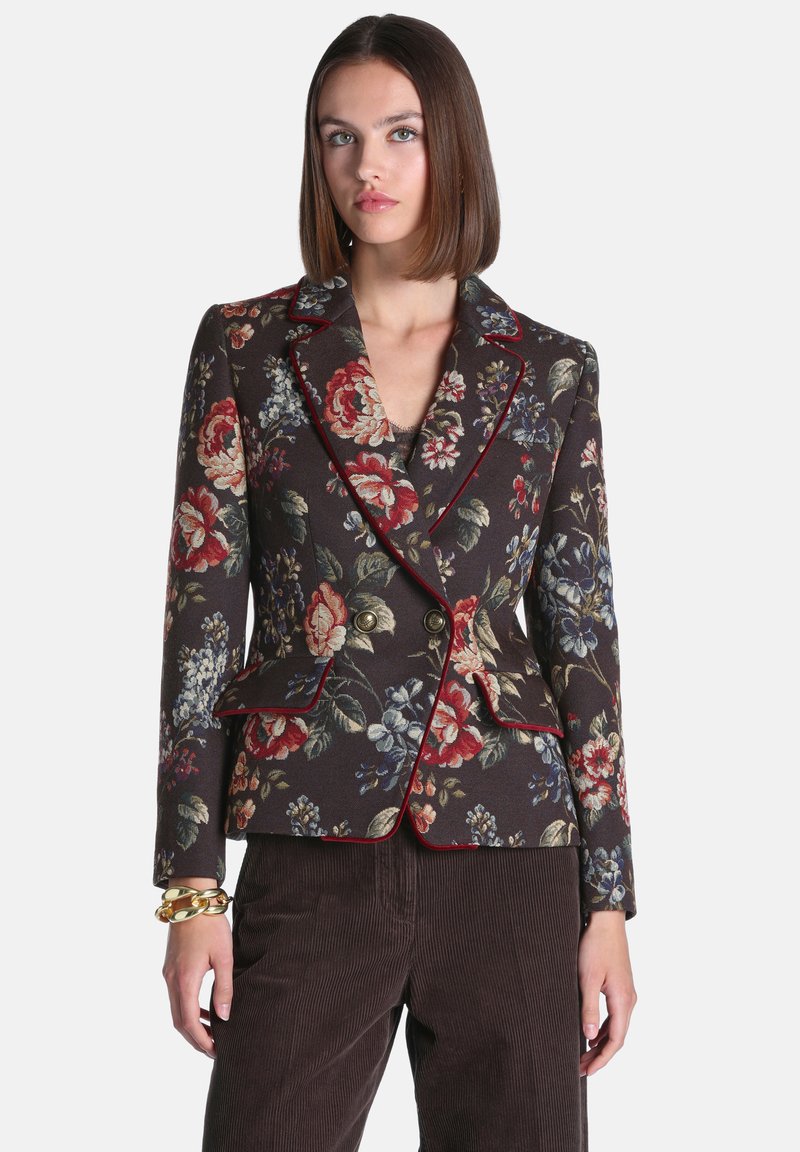Femme aux cheveux bruns et lisses portant un blazer fleuri foncé avec une bordure rouge et des boutons dorés, associé à un pantalon en velours marron et un bracelet en or.