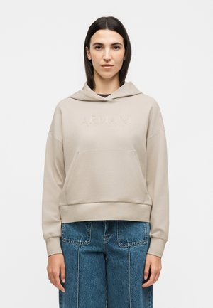 Frau trägt beigen Hoodie mit dezentem Armani-Logo und blaue High-Waist-Jeans, steht vor weißem, einfachem Hintergrund.