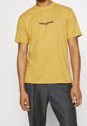 Mann trägt ein senfgelbes kurzärmeliges T-Shirt mit dem Schriftzug „THE KOOPLES PARIS“, kombiniert mit grauen plissierten Hosen vor weißem Hintergrund.