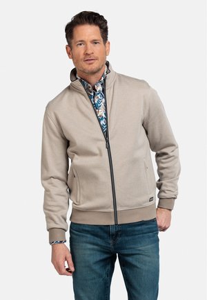 Veste zippée beige en tissu texturé avec des poignets côtelés, associée à une chemise à imprimé floral en dessous et à un jean en denim bleu.