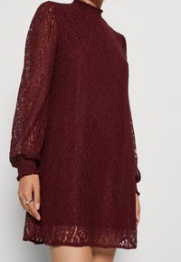 Vestido de encaje burdeos con mangas largas y fluidas, escote alto y un patrón floral texturizado. Presenta una silueta acampanada y puños con borde de pico.