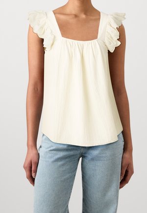 Vrouw draagt een crèmekleurige mouwloze top met geplooide eyelet-details op de schouders en lichtblauwe spijkerbroek.