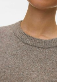 Gros plan sur le cou et la clavicule d'une personne portant un pull ras du cou en tricot doux, de couleur gris-brun, sur un fond blanc.