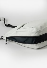Lichtgrijze tas met een strak ontwerp, voorzien van een contrasterende zwarte ritszak en een gestructureerd oppervlak. Inclusief verstelbare schouderband.